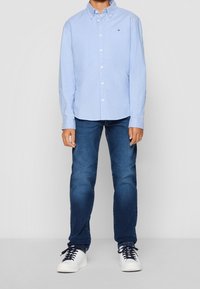 Junge trägt ein hellblaues Hemd mit Knöpfen, dunkelblaue Jeans und weiße Sneaker mit schwarzen Schnürsenkeln, steht vor einem schlichten Hintergrund.