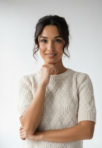 Kremowy sweter dzianinowy z teksturowanym wzorem w warkocze, krótkimi rękawami i prążkowanym dekoltem. Modelka pozuje zamyślona z ręką pod brodą.