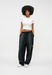 Λευκή κοντή μπλούζα με μαύρο λογότυπο, συνδυασμένη με μαύρα φόρμας που διαθέτουν στρογγυλή κόκκινη λεπτομέρεια. Το μοντέλο φορά λευκά αθλητικά παπούτσια.