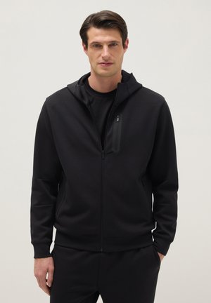 Man met een zwarte hoodie met rits en zwarte broek, staand met één hand in zijn zak tegen een effen lichte achtergrond.