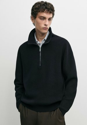 Jeune homme aux cheveux bouclés portant un pull noir à demi-fermeture éclair sur une chemise rayée, les mains dans les poches d’un pantalon sombre, sur un fond uni.