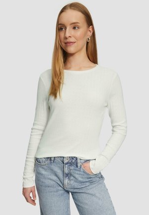 Leichtes, weißes, geripptes Langarmshirt mit rundem Ausschnitt und subtilen texturierten Muster, kombiniert mit hellblauen, hochgeschnittenen Jeans.