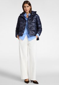 Granatowa pikowana kurtka puchowa z kapturem, z srebrnymi detalami i zatrzaskami, noszona na jasnoniebieskiej koszuli w paski i białych spodniach.