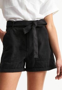 Black cotton shorts with a high waist, featuring a tie belt, rolled hems, and side pockets. Smooth texture and casual design.