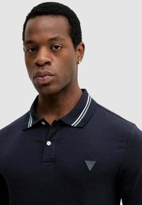 Granatowa koszulka polo z białym paskowanym kołnierzem, krótkimi rękawami i trójkątnym logo na lewej piersi. Gładka bawełniana tkanina.