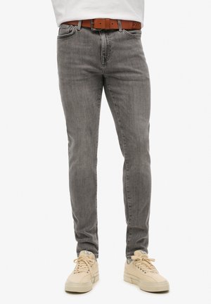 Grijze slim-fit spijkerbroek met een vervaagde textuur, met vijf zakken en een bruin leren riem, gecombineerd met lichtbeige sneakers.