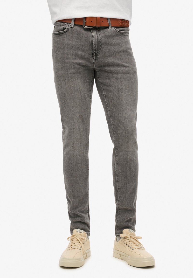 Szare slim-fit dżinsy z wyblakłą fakturą, wyposażone w pięć kieszeni i brązowy skórzany pasek, w połączeniu z jasnobeżowymi trampkami.