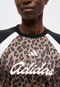 Άτομο με πουκάμισο Adidas με λεοπάρ μοτίβο, μαύρα και λευκά raglan μανίκια και μαύρο γιακά, εμφανίζεται από το λαιμό έως το στήθος.