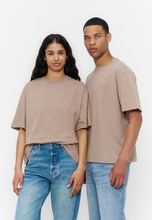 Zwei Personen stehen nebeneinander und tragen beige übergroße T-Shirts und blaue Jeans vor einem schlichten weißen Hintergrund.