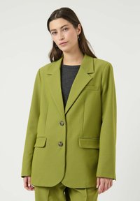 Blazer vert en tissu texturé, avec un col à revers cranté, deux poches avant et deux boutons. Porté sur un haut gris foncé.