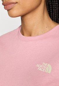 Ljust rosa t-shirt med rund halsringning, tillverkad av mjuk bomull, med en liten vit logotyp av "The North Face" på bröstet.