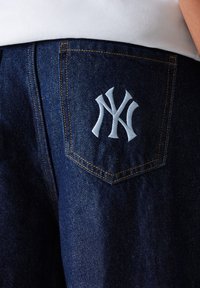Un jean en denim foncé présente une poche arrière avec un logo New York Yankees brodé en blanc. Les coutures sont orange, ajoutant un contraste au tissu.