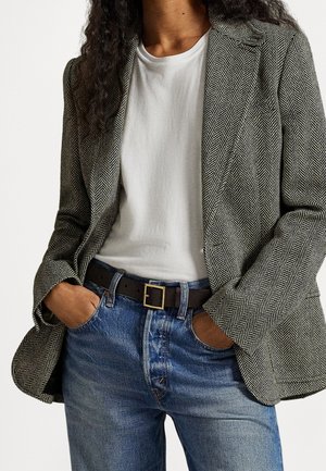 Personne les mains dans les poches, portant un blazer en chevrons noir et blanc, un t-shirt blanc, une ceinture noire et un jean en denim bleu.