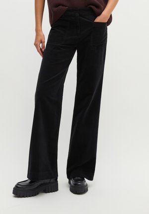 Personne portant un pantalon noir à jambes larges avec des poches avant, associé à des chaussures plateformes noires épaisses et un haut marron légèrement rentré.