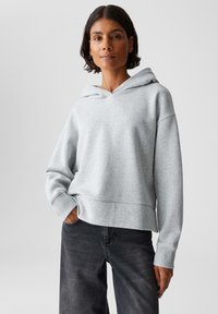 Una donna con i capelli corti e scuri indossa una felpa con cappuccio grigia chiara e jeans neri, in piedi con una mano in tasca su uno sfondo neutro.