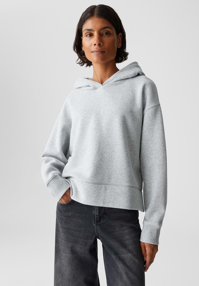 Una donna con i capelli corti e scuri indossa una felpa con cappuccio grigia chiara e jeans neri, in piedi con una mano in tasca su uno sfondo neutro.