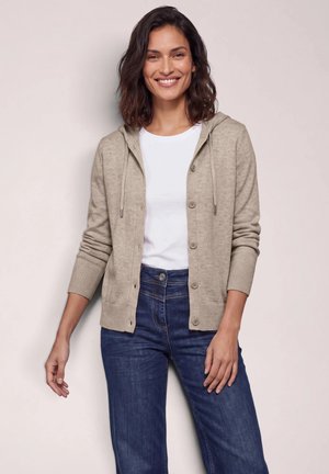 Mujer sonriente con una sudadera beige con botones sobre una camisa blanca y vaqueros azules, de pie contra un fondo liso.