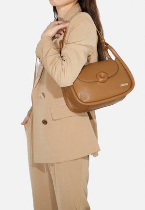 Borsa a tracolla in pelle marrone con chiusura a bottone rotondo, texture liscia e una lunga tracolla. Il modello indossa un completo beige su misura, mettendo in mostra la borsa.