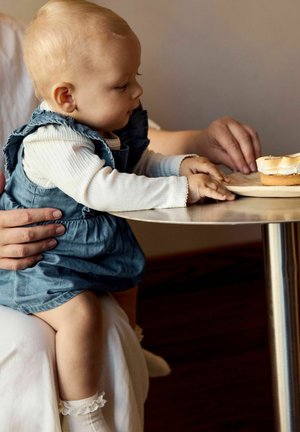 Batole v modrých šatech sahající po opékaném marshmallow dezertu položeném na dřevěném talíři na kulatém stole, poblíž dospělé ruce.