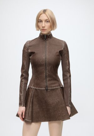 Donna con capelli biondi corti che indossa una giacca marrone aderente con zip e una gonna marrone abbinata su uno sfondo bianco uniforme.