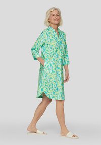 Leichtes, knielanges Kleid in Grün und Blau mit einem Blättermuster, mit hochrollbaren Ärmeln und einem knöpfbaren Ausschnitt. Glatte Stofftextur.