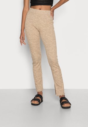 Kobieta w beżowych prążkowanych spodniach z bocznymi rozcięciami, czarnym krótki topie i czarnych sandałach stoi na tle jednolitego tła.