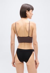 Γυναίκα με κοντά σκούρα μαλλιά, ντυμένη με καφέ τοπ με λεπτές τιράντες και μαύρο εσώρουχο με ψηλή μέση, όρθια με την πλάτη προς το μέρος μας, μπροστά σε απλό λευκό φόντο.