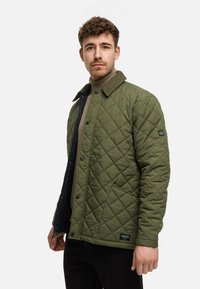 Grün gesteppte Jacke mit einem Cordkragen, vorderen Druckknopfverschlüssen und seitlichen Taschen, die ein strukturiertes Muster und ein Logopatch am Ärmel aufweist.