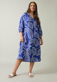 Vestido azul con patrón de hojas blancas, escote en V y mangas tres cuartos. La tela parece suave con un corte relajado, combinado con sandalias blancas.