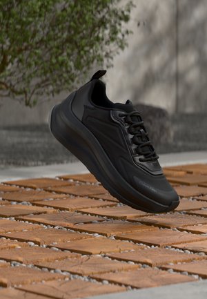 Μαύρο αθλητικό παπούτσι με χοντρή σόλα και κορδόνια που αιωρείται πάνω από ξύλινο πλακάκι, θαμπό φόντο εξωτερικού χώρου με πράσινο φύλλωμα και τοίχο.