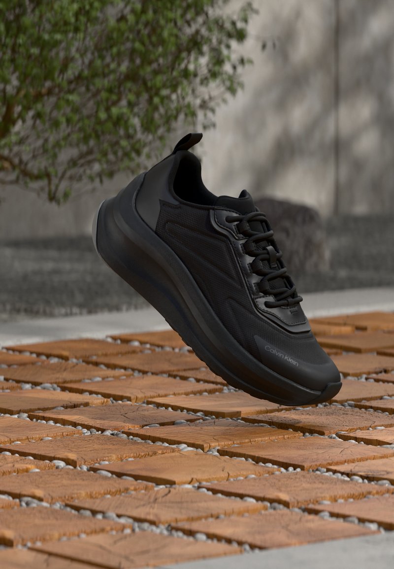 Μαύρο αθλητικό παπούτσι με χοντρή σόλα και κορδόνια που αιωρείται πάνω από ξύλινο πλακάκι, θαμπό φόντο εξωτερικού χώρου με πράσινο φύλλωμα και τοίχο.