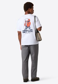 Mand iført hvid t-shirt med grafisk motiv af en gående bjørn og teksten "Gear Up. Get Out.", grå bukser og bærende en beige skuldertaske.
