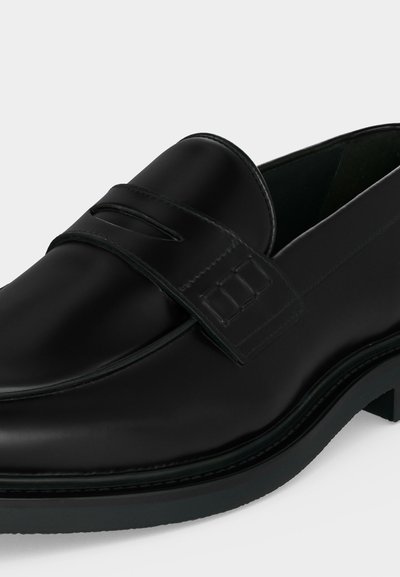 Μαύρο δερμάτινο μοκασίνι τύπου slip-on με στρογγυλεμένη μύτη, ραφές και διακοσμητικό λουράκι στο πάνω μέρος, εμφανιζόμενο από πλάγια γωνία.