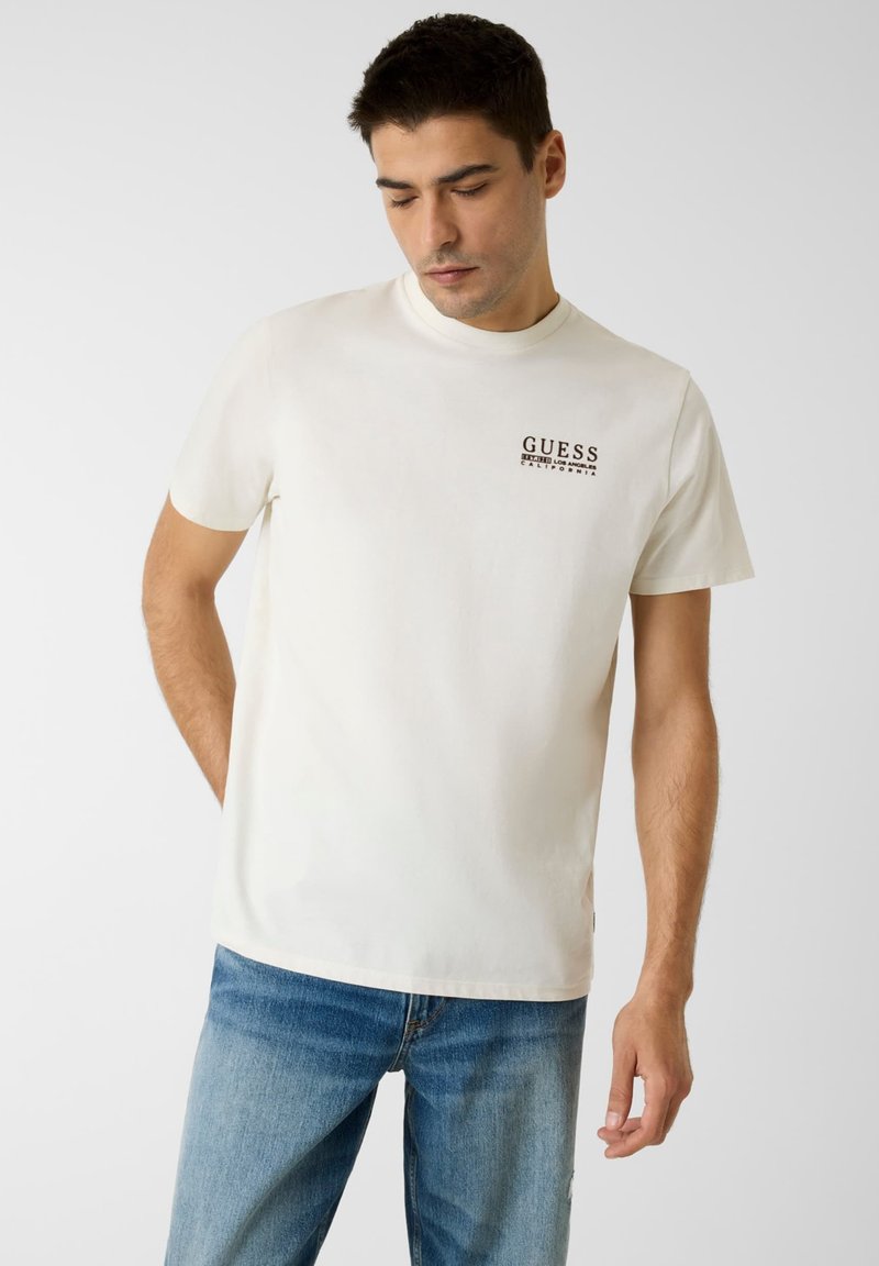 T-shirt en coton blanc avec un logo "GUESS" noir sur le côté gauche de la poitrine, accompagné d'un jean en denim bleu à coupe droite.