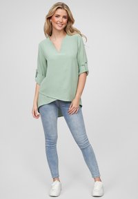 Maglia verde chiaro a maniche lunghe con scollatura a V e dettagli con orli arrotolati. Tessuto morbido con orlo asimmetrico, abbinata a jeans skinny blu chiaro.