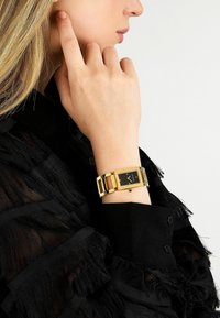 Orologio rettangolare tono oro con quadrante nero, cinturino a maglie e dettagli audaci, indossato su un polso contro una maglia nera con frange.