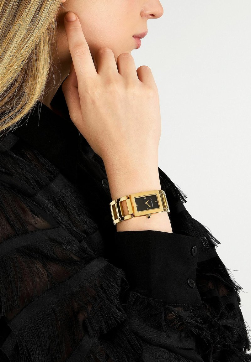 Orologio rettangolare tono oro con quadrante nero, cinturino a maglie e dettagli audaci, indossato su un polso contro una maglia nera con frange.