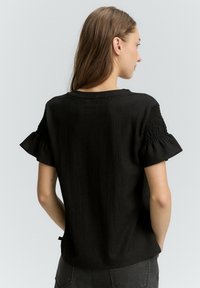 Sort tekstureret top med korte flæseærmer og sammenbragte detaljer. Klassisk rund halsudskæring. Letvægtsstof, enkelt design.