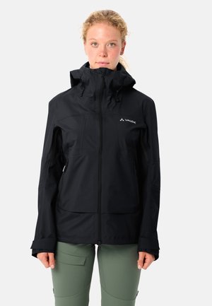 Junge Person trägt eine schwarze wasserdichte Jacke mit Kapuze, die hochgeschlossen ist, und hellgrüne Hosen, steht vor einem einfarbigen hellen Hintergrund.