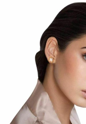 Donna con capelli scuri raccolti indietro che indossa piccoli orecchini a lobo a forma di trifoglio in oro e perle e una leggera camicia di raso su sfondo bianco.