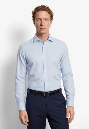 Uomo con capelli ricci che indossa una camicia azzurra con bottoni, pantaloni blu scuro e una cintura nera, in piedi davanti a uno sfondo bianco.