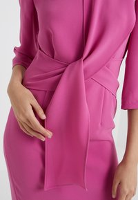 Fuchsiové šaty s hladkým, lehkým materiálem. Mají detail na zavázání v oblasti pasu a rukávy po třičtvrtě, což ukazuje na elegantní, přiléhavý siluetu.