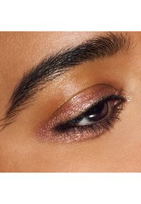 Ingrandimento di un occhio chiuso con ombretto rosa oro lucido e sopracciglio scuro, ben curato e spesso, su pelle liscia.