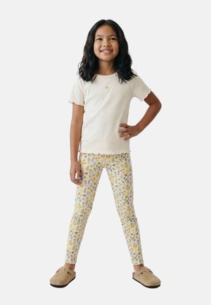 Jeune fille aux cheveux noirs mi-longs posant avec le sourire, portant un t-shirt blanc à manches courtes, des leggings à fleurs et des chaussures beiges sans lacets.