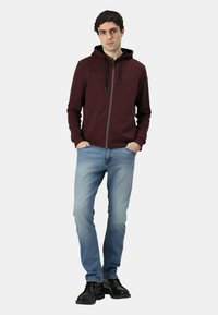 Felpa con cappuccio bordeaux con zip e coulisse nere; indossata sopra jeans azzurri e scarpe nere. Tessuto morbido, design semplice con dettagli minimi.