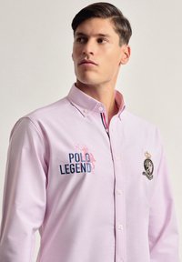 Camisa de botón de color rosa claro hecha de algodón. Presenta bordado "POLO LEGEND" en azul, un parche con el logo y acentos a rayas en el collar.