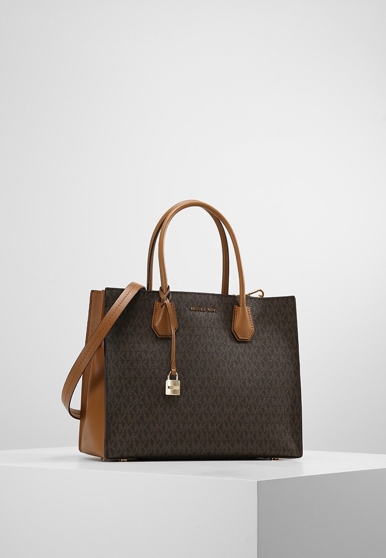 Sac à main Michael Kors marron et sombre à motifs, avec deux poignées, une bandoulière et un petit cadenas doré suspendu, sur un socle blanc.