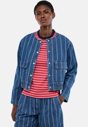 Femme portant une veste et un pantalon en denim bleu à fines rayures, avec une chemise rayée rouge et blanche, debout avec une main dans la poche.