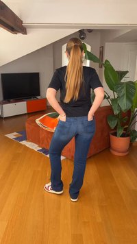 Γυναίκα με μακριά αλογοουρά που φοράει μαύρο πουκάμισο και μπλε τζιν στεκόταν με τα χέρια στις πίσω τσέπες σε ένα σαλόνι δίπλα σε ένα μεγάλο φυτό.