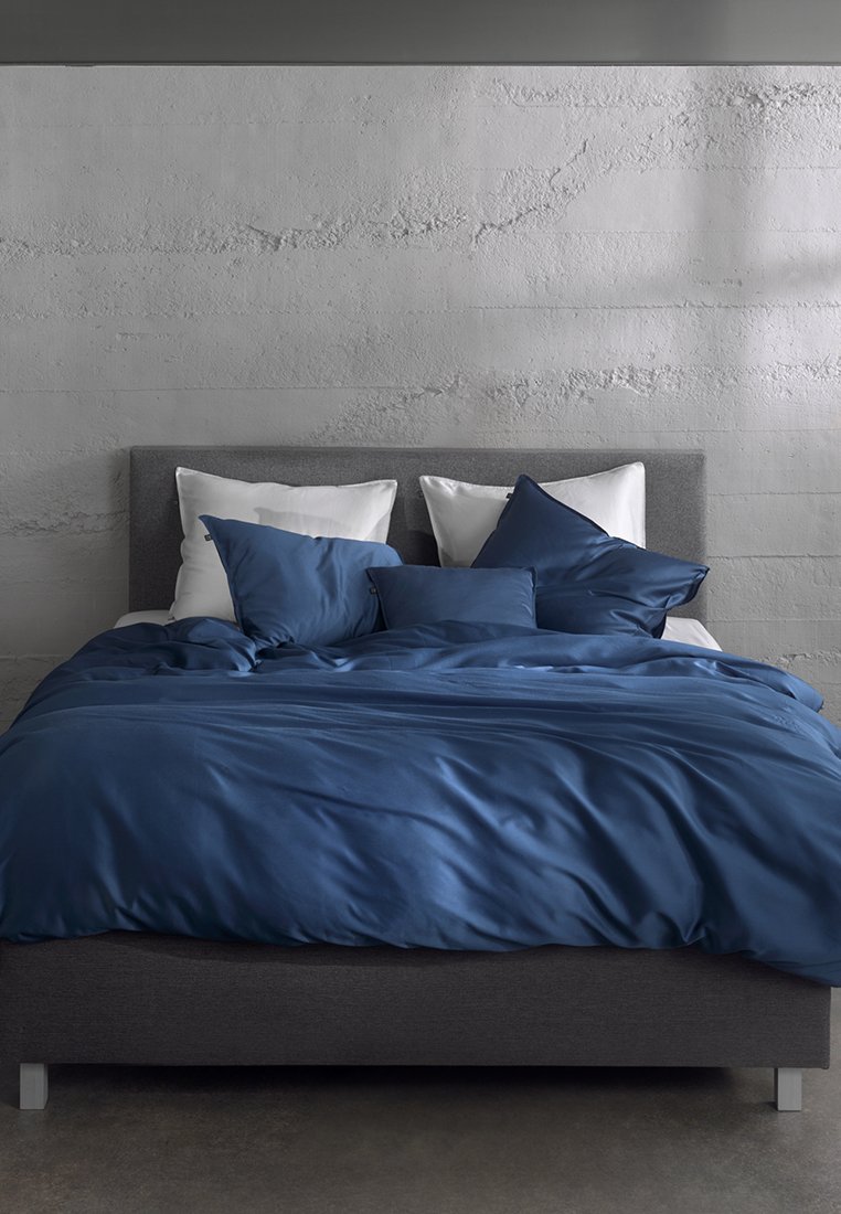 Dvoulůžková postel s šedou čalouněnou čelní deskou, bílými polštáři a modrým povlakem na peřinu a polštáři proti texturované šedé zdi.